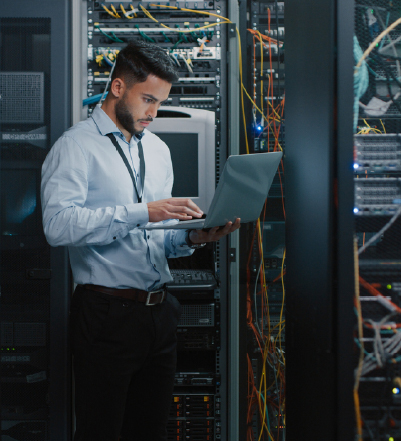 Data center technician working on a laptop beside server racks and network equipment.