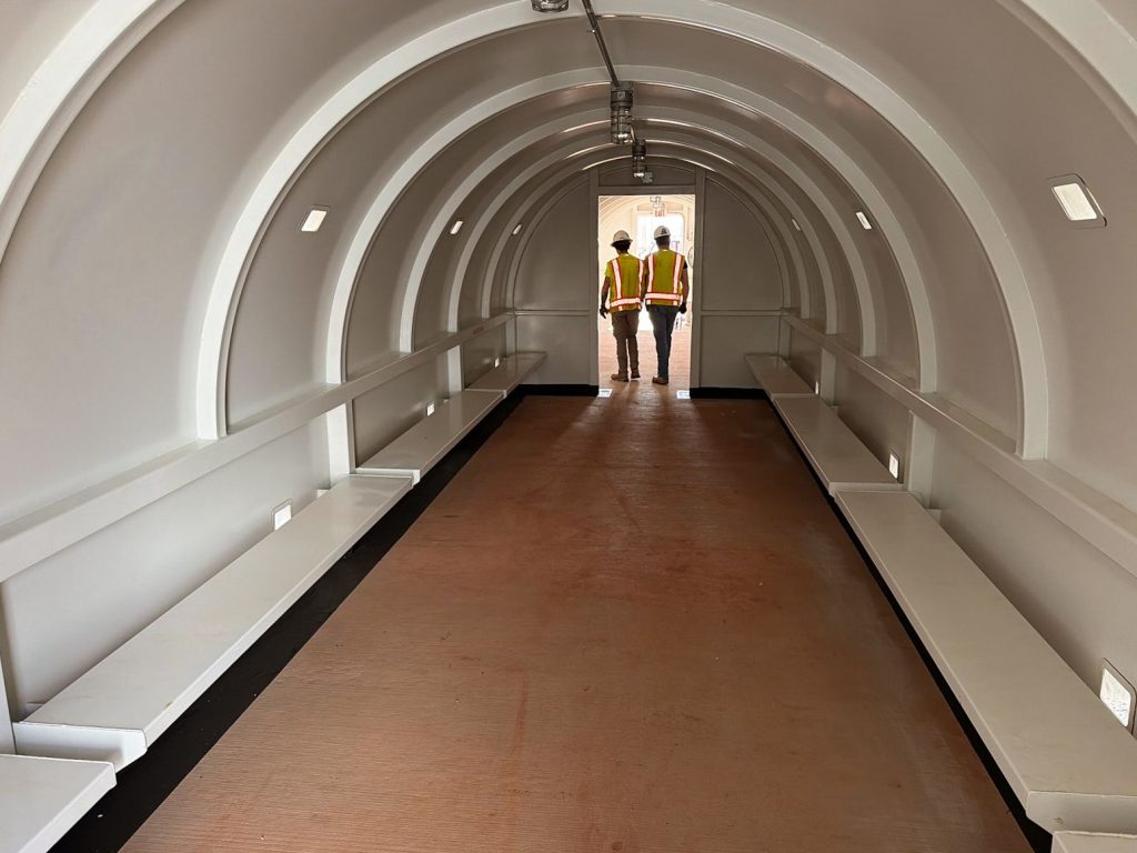 Interior of a commercial tornado shelter with built-in bench seating and two workers standing near the entrance.