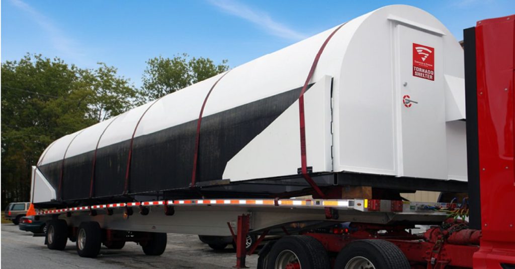Prefabricated commercial tornado shelter secured to a flatbed trailer for delivery to an installation site.
